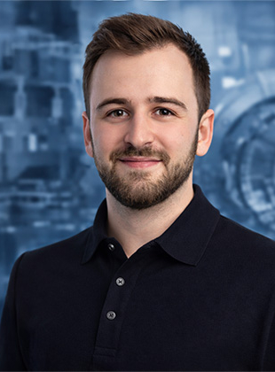 Bild: Das Bild zeigt Projektleiter Julian Schaufler. Julian ist ein junger Mann mit braunen Haaren, braunen Augen und einem dunklen Bart, im Polohemd, der auf dem Bild freundlich lächelt.